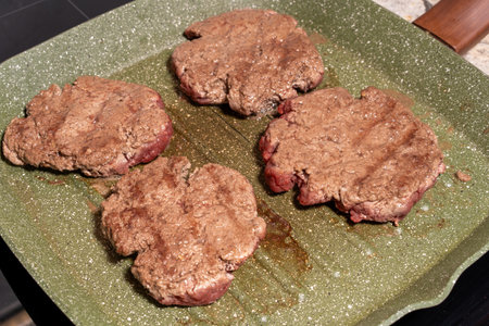 Four hamburger patties cooking on a green speckled griddle. The patties show visible browning and texture, surrounded by grease, emphasizing the sizzling preparation process.の写真素材