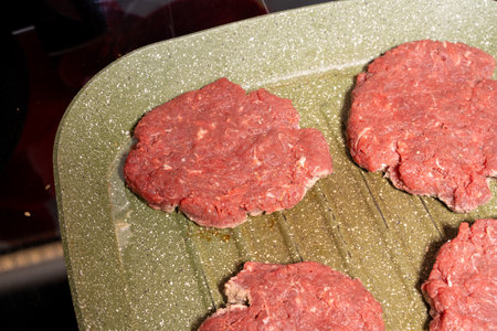 Four raw hamburger patties made of ground beef, placed on a green, speckled grill pan with ridged texture. The uncooked patties are ready for grilling, highlighting preparation for a delicious meal.の写真素材