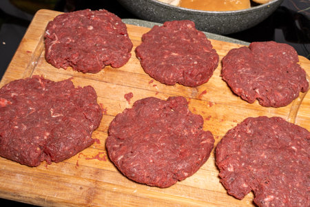 Six uncooked hamburger patties made of ground meat are placed on a wooden cutting board. In the background, hamburger buns are being toasted in a frying pan, illustrating burger preparation.の写真素材