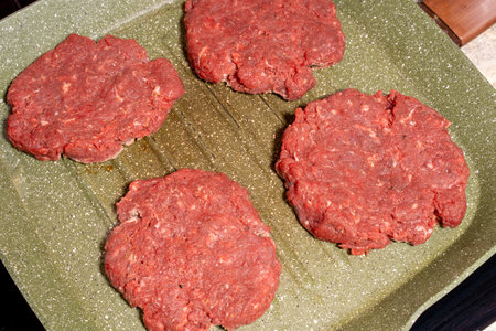 Four raw hamburger patties made of ground beef, placed on a green, speckled grill pan with ridged texture. The uncooked patties are ready for grilling, highlighting preparation for a delicious meal.の写真素材