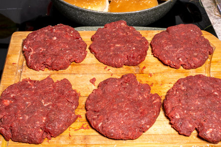 Six uncooked hamburger patties made of ground meat are placed on a wooden cutting board. In the background, hamburger buns are being toasted in a frying pan, illustrating burger preparation.の写真素材