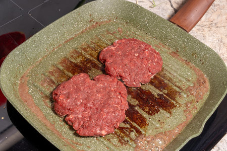 Two raw hamburger patties rest on a speckled green grill pan with visible grease marks. The coarse-textured patties highlight the preparation stage of homemade burgers, ready for grilling.の写真素材