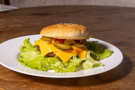 A cheeseburger on a white plate placed on a wooden surface. The burger features a sesame seed bun, beef patty, melted cheese, and green lettuce, showcasing a fresh homemade classic.の写真素材