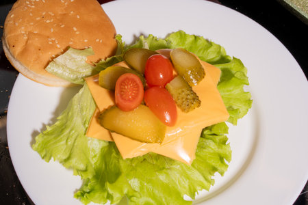 An open-faced burger on a white plate. Ingredients include a sesame seed bun, lettuce, melted cheese, pickles, and cherry tomatoes, showcasing a vibrant burger assembly presentation.の写真素材