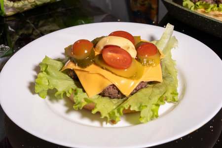 An open-faced burger on a white plate, featuring a beef patty with melted yellow cheese, crisp lettuce, pickles, cherry tomatoes, and mayonnaise. The setup highlights a vibrant burger assembly.の写真素材