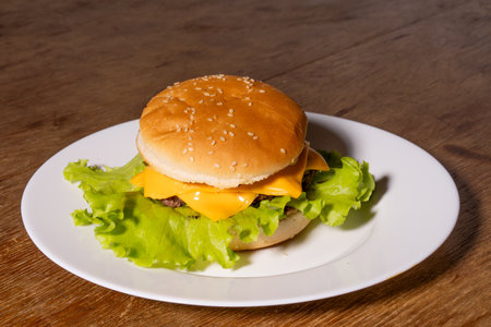 A cheeseburger on a white plate placed on a wooden surface. The burger features a sesame seed bun, beef patty, melted cheese, and green lettuce, showcasing a fresh homemade classic.の写真素材