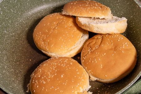 Four sesame seed hamburger buns in a speckled non-stick frying pan. One bun is partially cut, showing burger preparation. Overlapping arrangement adds a casual, homemade cooking feel.の写真素材