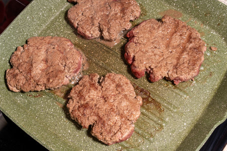 Four hamburger patties cooking on a green speckled griddle. The patties show visible browning and texture, surrounded by grease, emphasizing the sizzling preparation process.の写真素材