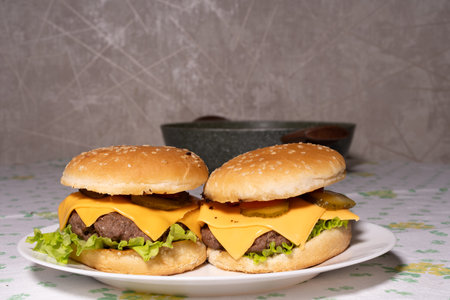 Two well-assembled cheeseburgers with sesame buns, beef patties, cheddar cheese, lettuce, pickles, and tomato on a white plate, surrounded by kitchen items in the background.の写真素材