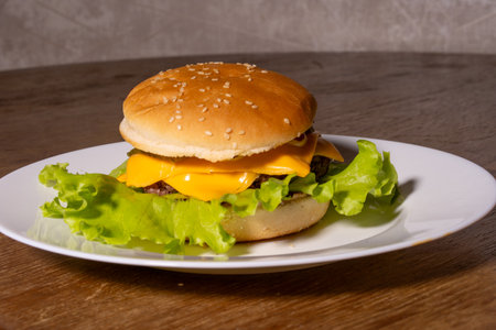 A cheeseburger on a white plate placed on a wooden surface. The burger features a sesame seed bun, beef patty, melted cheese, and green lettuce, showcasing a fresh homemade classic.の写真素材