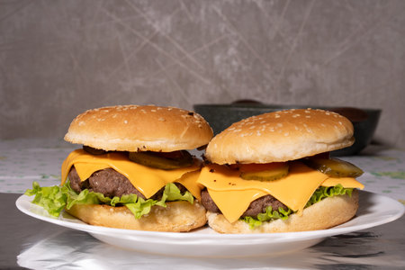 Two well-assembled cheeseburgers with sesame buns, beef patties, cheddar cheese, lettuce, pickles, and tomato on a white plate, surrounded by kitchen items in the background.の写真素材