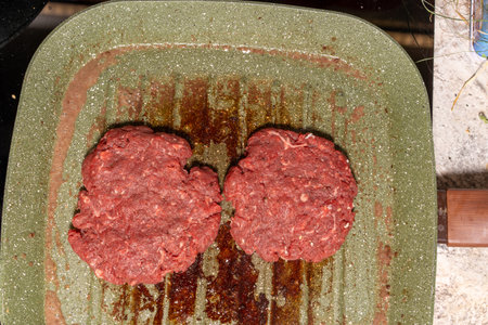 Two raw hamburger patties rest on a speckled green grill pan with visible grease marks. The coarse-textured patties highlight the preparation stage of homemade burgers, ready for grilling.の写真素材