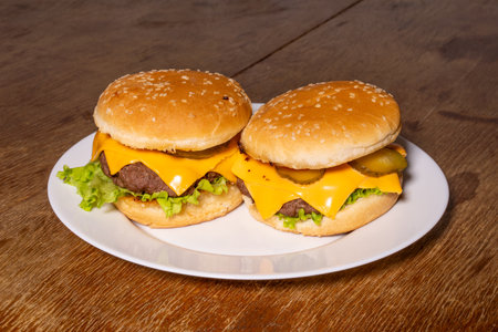 Two cheeseburgers on a white plate, featuring sesame seed buns, beef patties, melted cheese slices, pickles, and crisp lettuce, set on a rustic wooden surface.の写真素材