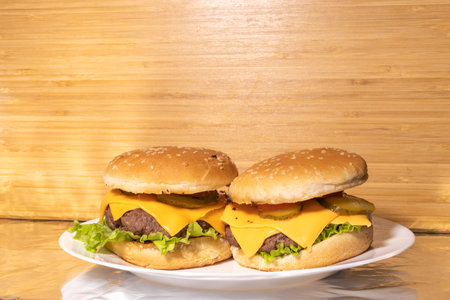 Two cheeseburgers with sesame seed buns, beef patties, cheese, pickles, tomato slices, and lettuce are served on a white plate. A rustic wooden surface forms the background.の写真素材