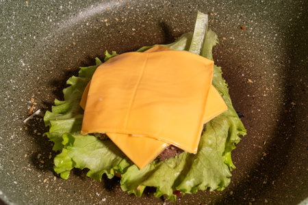 A frying pan holds a hamburger patty topped with two melted slices of cheese resting on crisp lettuce, highlighting a key step in burger assembly during cooking.の写真素材