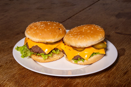 Two cheeseburgers on a white plate, featuring sesame seed buns, beef patties, melted cheese slices, pickles, and crisp lettuce, set on a rustic wooden surface.の写真素材
