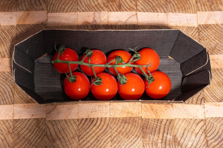 Red cherry tomatoes still attached to a vine, resting in a cardboard container on a patterned wooden cutting board. Fresh, vibrant, and ideal for culinary or rustic food scenes.の写真素材