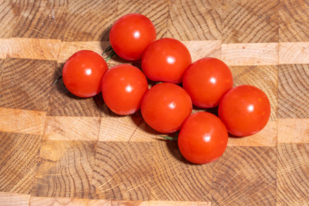 Eight red cherry tomatoes on a green vine rest on a wooden cutting board with alternating light and dark wood grain patterns, showing freshness and rustic simplicity.の写真素材