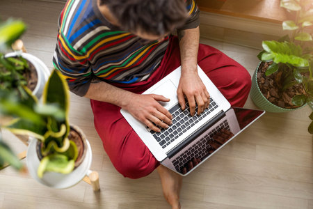 Young man working at home with laptop, sitting on the floor surrounded by houseplantsの写真素材