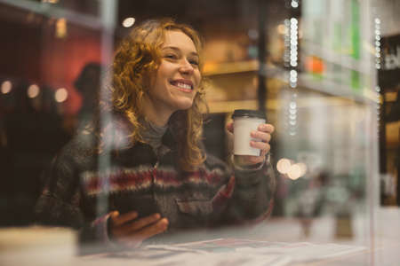 Smiling young woman with cup of coffee behind window in a cafeの写真素材