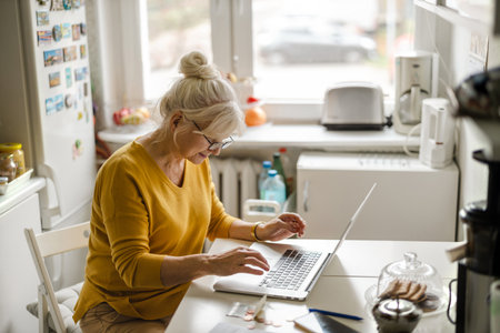 Senior woman using laptop in kitchen at home. Senior woman using computer at home.