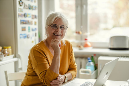 Portrait of smiling senior woman looking at camera while working on laptop in the kitchenの写真素材