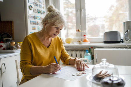 Mature woman writing on paper while sitting at the table in the kitchenの写真素材