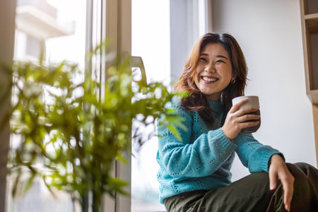 Portrait of a smiling asian woman sitting on the windowsill and holding a cup of coffeeの写真素材