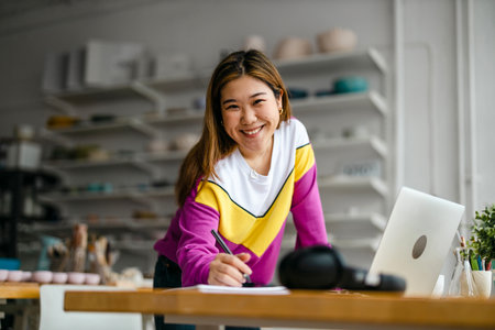 Beautiful young asian woman smiling while working with laptop in workshopの写真素材