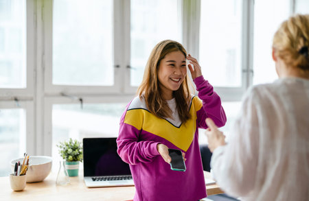 Two young businesswomen talking on mobile phone while sitting at desk in officeの写真素材
