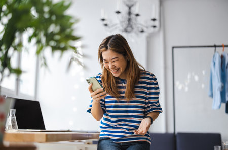 happy young asian woman using smartphone while sitting at workplace in officeの写真素材
