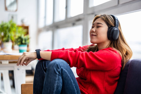Young asian woman listening to music with headphones while sitting on sofa at homeの写真素材