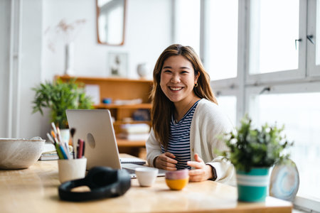 Smiling young asian woman using laptop while sitting at table in officeの写真素材
