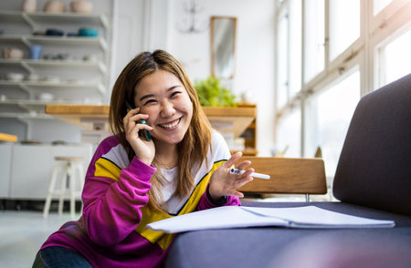 Smiling asian woman talking on the phone and writing notes at homeの写真素材
