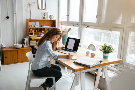Beautiful young businesswoman working on laptop while sitting at desk in officeの写真素材