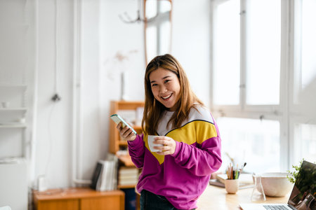 Portrait of a smiling young woman using mobile phone while drinking coffee at homeの写真素材