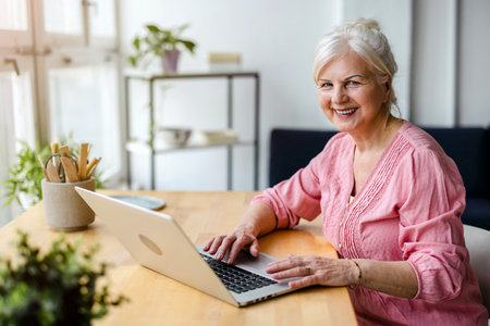 Portrait of smiling senior woman using laptop while sitting at table at homeの写真素材