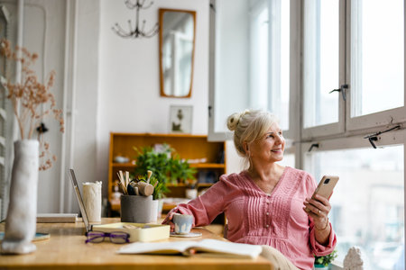 Smiling senior woman using mobile phone while sitting at table in creative officeの写真素材