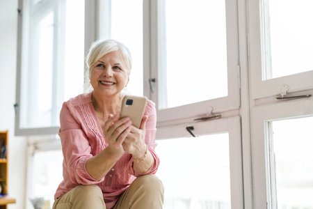 Portrait of smiling senior woman using mobile phone while sitting at homeの写真素材