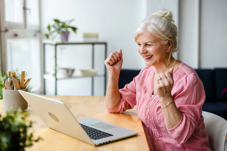 Cheerful senior woman celebrating success in front of laptop at homeの写真素材