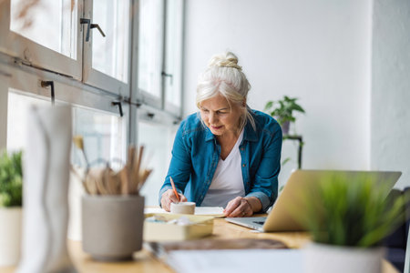 senior woman drawing with pencils while sitting at table in officeの写真素材