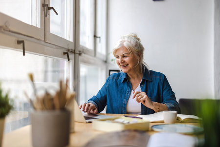 Smiling mature woman using laptop while sitting at table in creative officeの写真素材