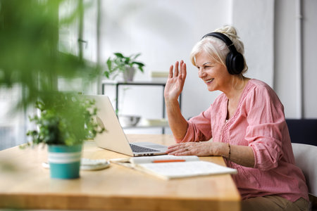 senior woman in headphones using laptop and waving hand while sitting at homeの写真素材