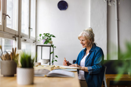 smiling mature businesswoman writing in notebook while sitting at table in officeの写真素材