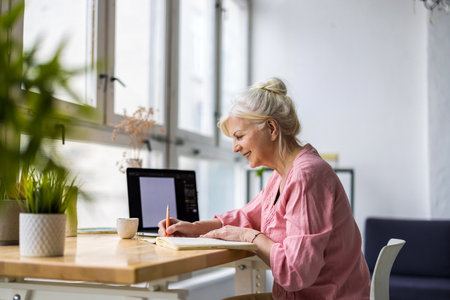 senior woman writing in notebook while sitting at table with laptop at homeの写真素材
