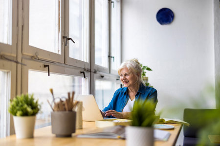 smiling senior businesswoman using laptop at office desk in front of windowの写真素材