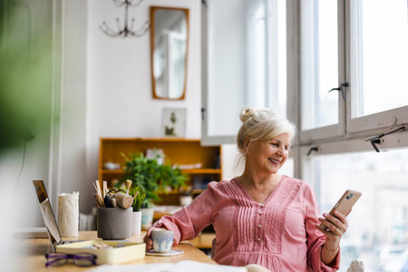 Smiling mature businesswoman using mobile phone while sitting at desk in officeの写真素材