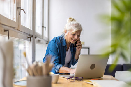 Smiling businesswoman talking on mobile phone while using laptop in officeの写真素材