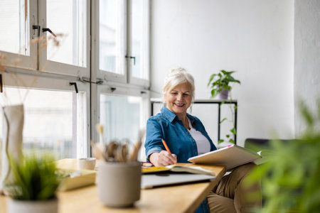 smiling mature woman writing in notebook while sitting at table in officeの写真素材