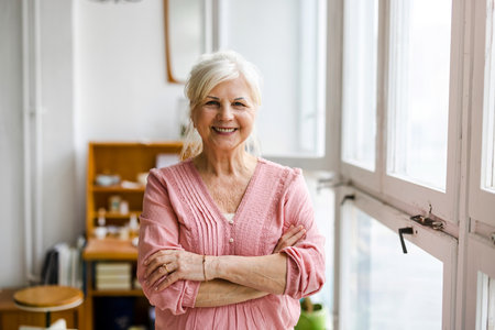 Portrait of smiling senior woman standing with arms crossed in kitchen at homeの写真素材